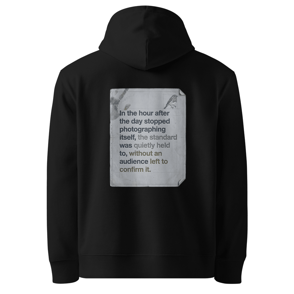 Quietly Held hoodie, black — back view of torn paper slogan plate with robin, reading "In the hour after the day stopped photographing itself, the standard was quietly held to, without an audience left to confirm it." — Ricki Robin
